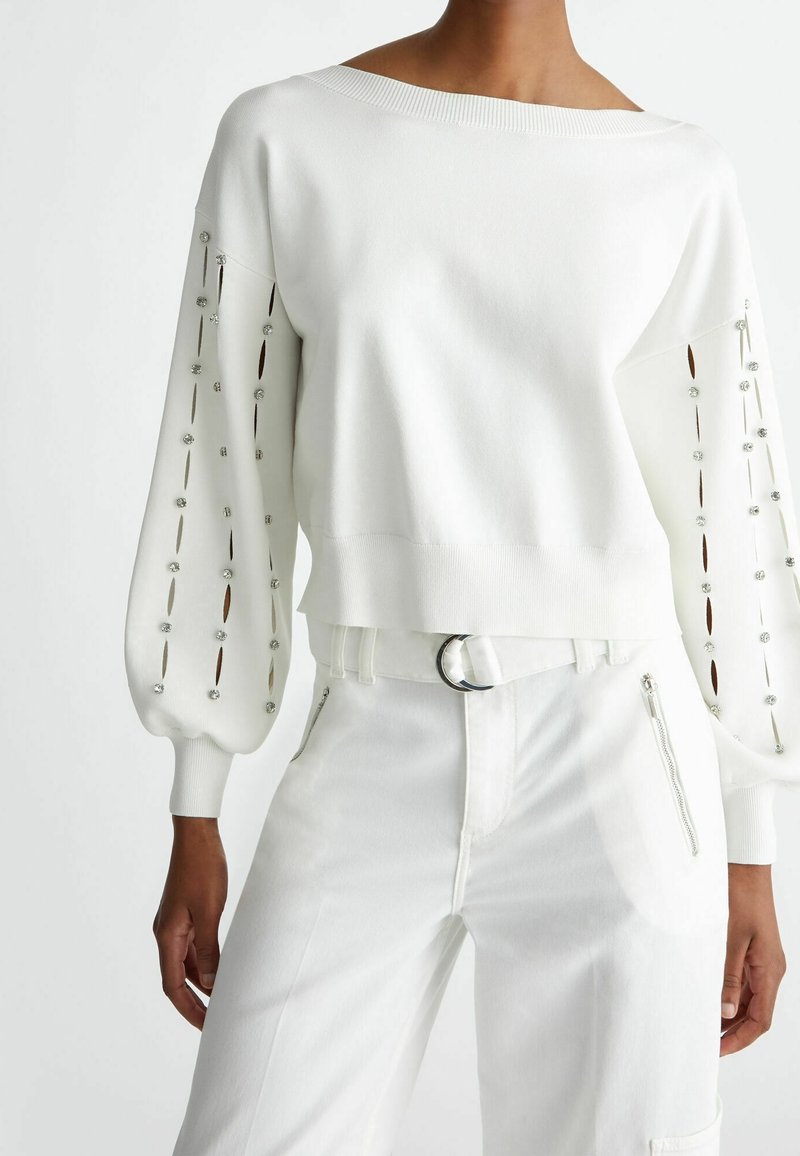 Femme portant un pull blanc épaules dénudées avec des détails en strass et fentes sur les manches bouffantes, associé à un pantalon blanc et une ceinture.