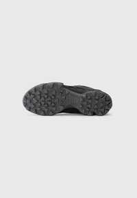 Suela de una zapatilla deportiva negra con patrones de tracción en forma de triángulos, mostrada sobre un fondo plano de color gris claro.