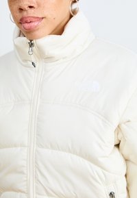 Beige puffjakke med høy krage, glidelås og subtil logo. Har en quiltet tekstur og glatt overflate. Sølvfarget detaljer.