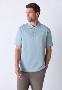 Polo à manches courtes en mélange de coton bleu clair avec un motif texturé, une patte à trois boutons et un col classique. Associé à un pantalon beige.