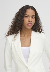 Kvinde med langt krøllet hår iført en hvid blazer over en hvid top, der ser selvsikkert på kameraet mod en ensartet lys baggrund.