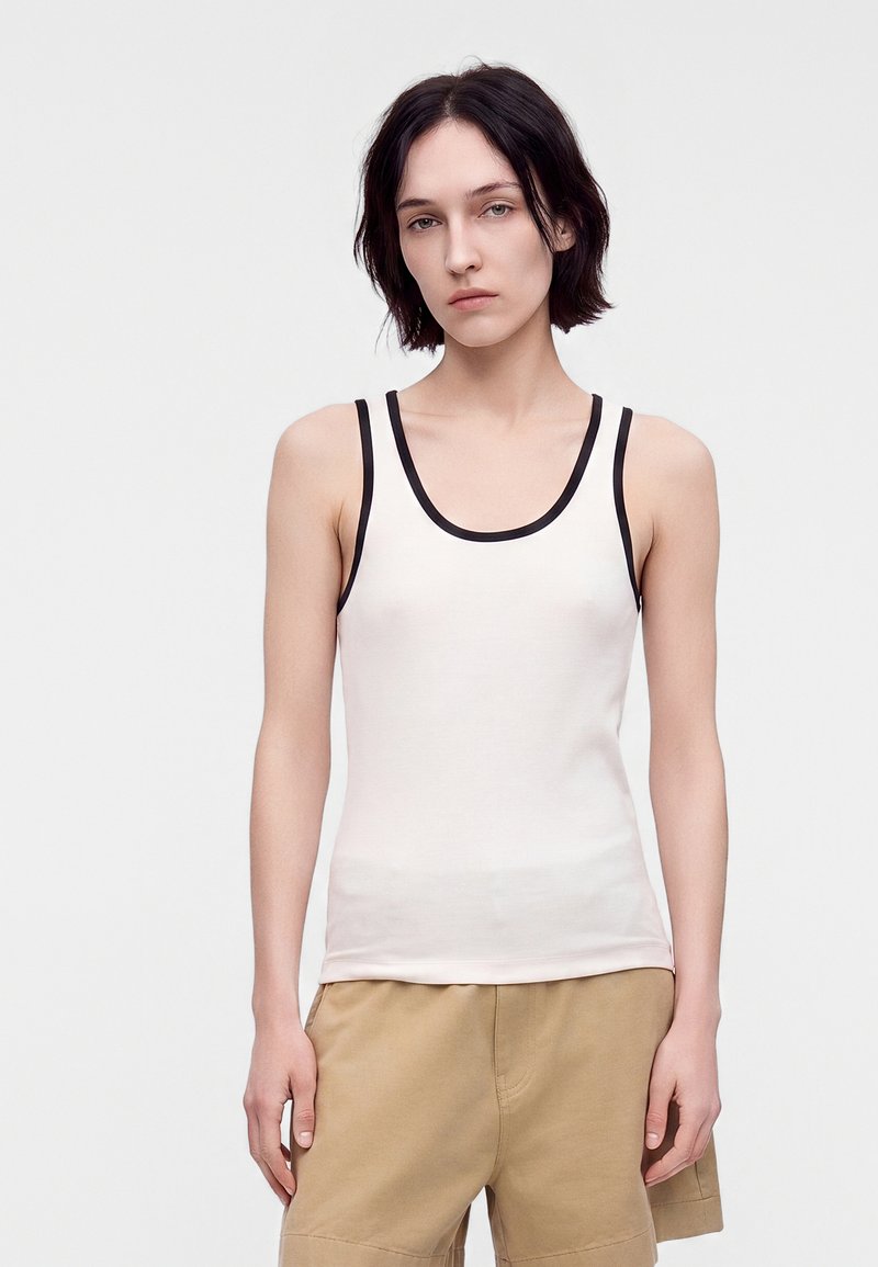 Jeune personne aux cheveux courts et foncés portant un débardeur blanc avec bordure noire et un short beige, debout devant un fond clair uni.