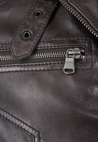 Braune Lederjacke mit einem Reißverschluss, metalischen Beschlägen und sichtbaren Nähdetails. Die Textur ist leicht genoppt.