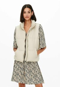 Gilet rembourré beige clair avec un col haut, fermeture éclair et deux poches latérales, porté par-dessus une robe à motifs à manches courtes.