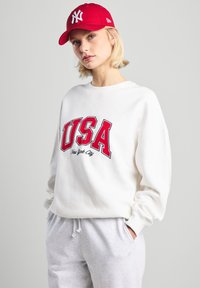 Felpa bianca con "USA" e "New York City" in rosso, abbinata a pantaloni della tuta grigi chiari e un cappello rosso con logo bianco.