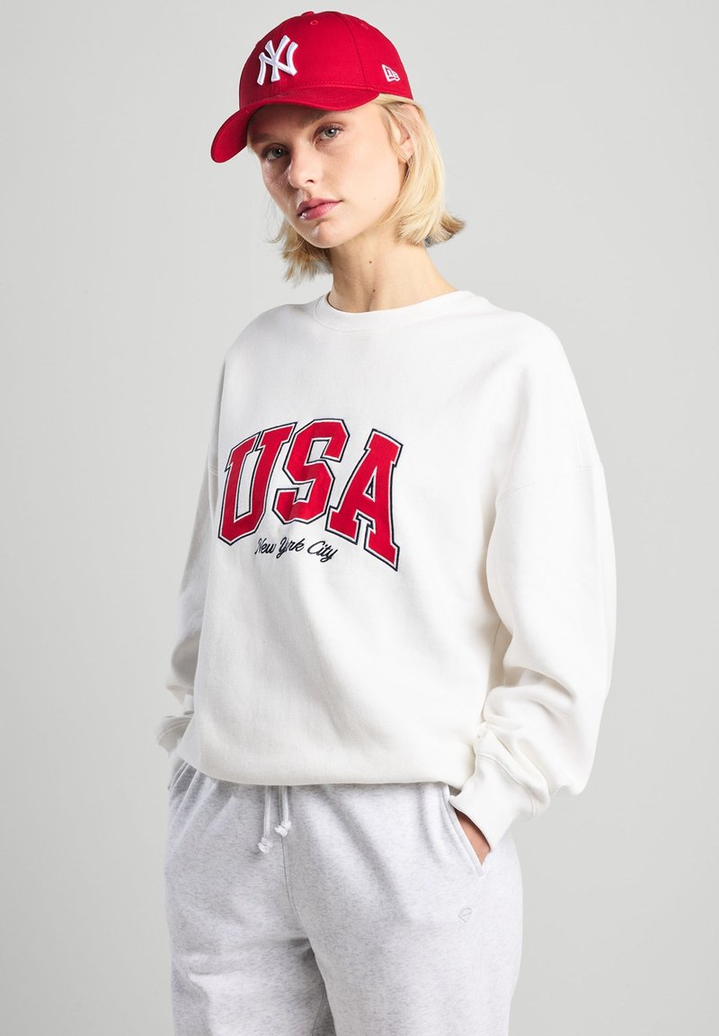 Felpa bianca con "USA" e "New York City" in rosso, abbinata a pantaloni della tuta grigi chiari e un cappello rosso con logo bianco.