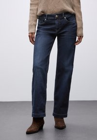 Jeans de mezclilla azul oscuro con corte recto, costuras visibles y un diseño clásico de cinco bolsillos. Llevados con un suéter de punto beige y botines marrones.