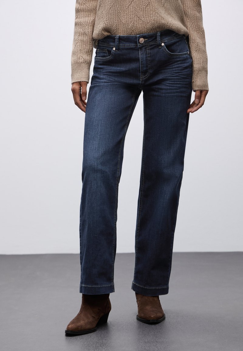 Jeans de mezclilla azul oscuro con corte recto, costuras visibles y un diseño clásico de cinco bolsillos. Llevados con un suéter de punto beige y botines marrones.