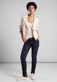 Veste beige clair, jeans noirs et sneakers blancs. La veste a une coupe décontractée avec de grands revers et deux poches avant.