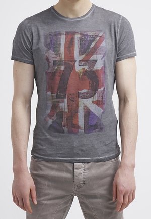 Mężczyzna w wyblakłej szarej koszulce z rozdartą flagą Wielkiej Brytanii i dużą liczbą 73 nadrukowaną z przodu.