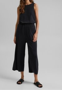 Combinaison noire sans manches, taille élastique et larges ouvertures aux jambes. Portée avec des sandales plates noires. Texture de tissu lisse.