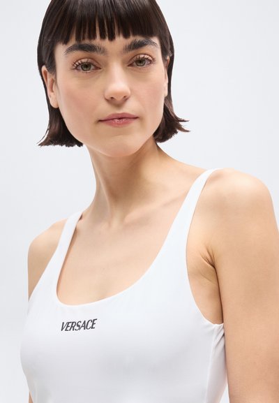 Γυναίκα με κοντά σκουρόχρωμα μαλλιά που φοράει λευκό τοπ με το λογότυπο «VERSACE», κοιτάζοντας ελαφρώς προς τα δεξιά μπροστά σε απλό ανοιχτό φόντο.
