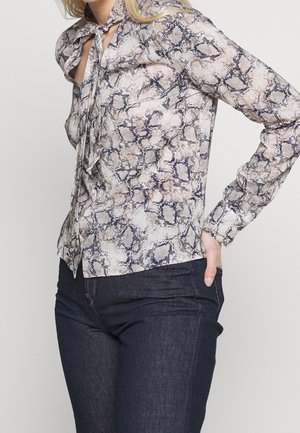 Donna che indossa una blouse a maniche lunghe con motivo pelle di serpente e dettaglio con fiocco al collo e jeans denim blu scuro, posando con la mano sui fianchi.