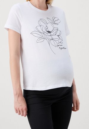 Biała koszula z krótkim rękawem z czarną grafiką kwiatową i napisem „Rośnijmy razem”, noszona przez osobę w czarnych spodniach na jednolitym tle.