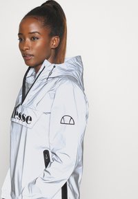 Hellblaues Kapuzenwindbreaker aus synthetischem Material mit einem Logo-Patch auf der Vorderseite, seitlichen Reißverschlusstaschen und schwarzen Akzentdetails.