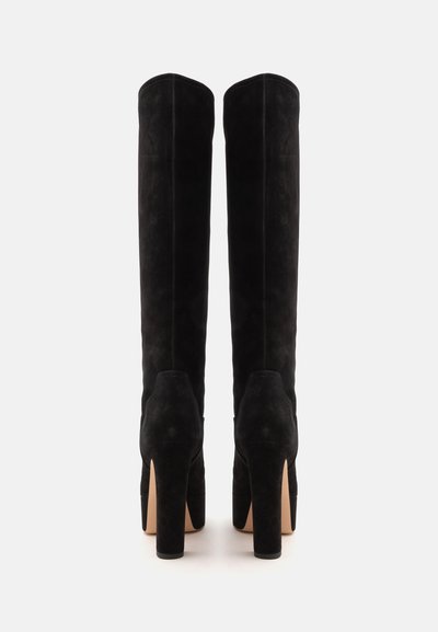 Paire de bottes hautes en daim noir à talons hauts vues de dos sur un fond blanc uni.