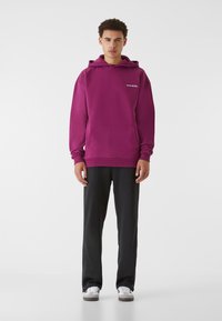 Felpa viola in tessuto morbido, con tasca a marsupio e logo bianco sul petto, abbinata a pantaloni neri e sneakers grigie.
