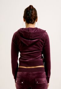 Felpa in velluto viola con cappuccio e polsini aderenti, abbinata a pantaloni in velluto dello stesso colore con tasche frontali dotate di chiusure a bottone.