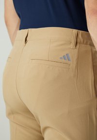 Pantalon beige en tissu léger, avec une coupe ajustée, deux poches arrière et un logo Adidas gris sur le côté droit.