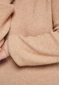 Pull en tricot beige avec texture côtelée, présentant des manches larges et un ourlet ajusté. Le matériau semble doux et chaud.