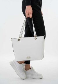 Witte tote bag met een getextureerd oppervlak, zwarte randen en gouden hardware. Heeft dubbele handvatten. Gepaard met witte sneakers en zwarte broek.