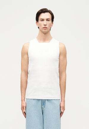 Fiatal férfi rövid sötét hajjal, ujjatlan fehér felsőt és világoskék farmert viselve, egy egyszínű világos háttér előtt áll.