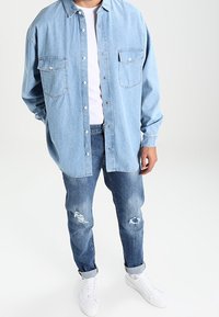 Mężczyzna w jasnoniebieskiej, oversize'owej koszuli dżinsowej noszonej na białej koszulce, w podartych niebieskich dżinsach z podwiniętymi mankietami oraz białych trampkach.