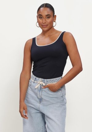 Woman with hair pulled back wears black sleeveless top with white trim and light blue high-waist jeans, standing with one hand in pocket.