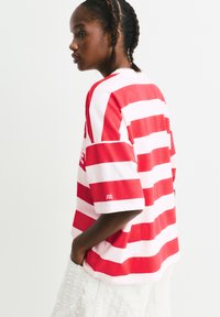 Junge Person mit geflochtenem Haar, die ein lockeres rot-weiß gestreiftes Shirt und strukturierte weiße Shorts trägt, seitlich blickend mit den Händen in den Taschen.