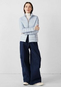 Chaqueta acolchada azul claro con cuello alto, cremallera frontal y mangas largas. Combinada con pantalones anchos azul oscuro y zapatillas blancas.