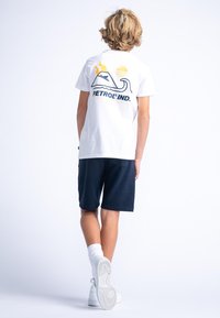 Biała koszulka z graficznym wzorem przedstawiającym fale, góry i słońce w kolorach niebieskim, żółtym i pomarańczowym. Połączona z granatowymi szortami i białymi trampkami.