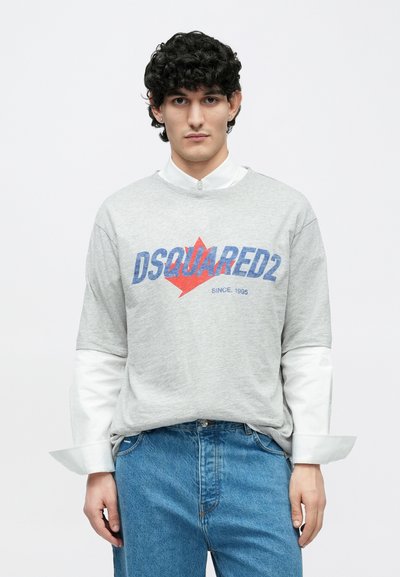 Pelēka kokvilnas T-krekls ar sarkanu un zilu logotipu, valkāts virs baltas pogu krekla. Piemērots valkāšanai kopā ar zilām džinsa biksēm.