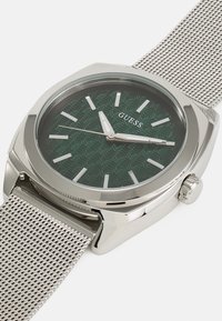 Reloj de acero inoxidable plateado con un dial texturizado verde, marcadores de horas blancos y una correa de malla. Presenta un bisel pulido y una corona.