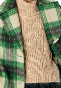 Cappotto di lana a quadri verdi e grigi sovrapposto a un maglione a collo alto beige lavorato a maglia con un motivo testurizzato. Il cappotto presenta dettagli con bottoni.