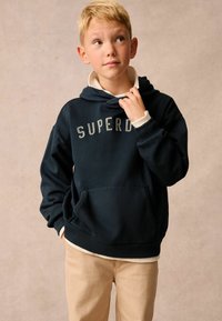 Sudadera con capucha azul marino con el logotipo "SUPERDRY", bolsillo frontal y puños acanalados, superpuesta a una camisa de color claro, combinada con pantalones beige.