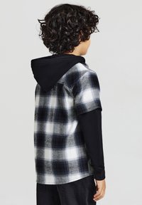 Camicia a maniche corte a quadri in bianco e nero con accenti grigi, indossata sopra una maglietta nera a maniche lunghe. Presenta un cappuccio nero.