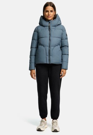 Blauwe pufferjack met capuchon, met een gewatteerd ontwerp, voorrits en zijzakken; gedragen met zwarte joggers en sneakers.
