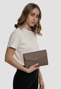 Borsa a tracolla in pelle marrone con finitura testurizzata e chiusura a pattina. Logo dorato visibile sul davanti. Modello che tiene la borsa contro uno sfondo neutro.