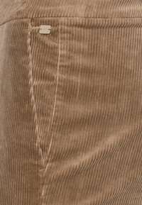 Tessuto di velluto a coste marrone con righe verticali e cuciture visibili, caratterizzato da un piccolo tag metallico rettangolare del marchio vicino al bordo della tasca.