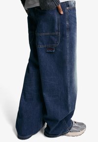 Hlače od denima s širokim nogavicama u tamnoplavoj boji, s prednjim i stražnjim džepovima te oznakom marke na stražnjem dijelu. Materijal od pamuka s izblijedjelom teksturom.