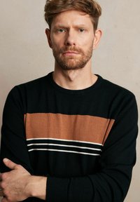 Czarny sweter z poziomym brązowym paskiem i dwoma białymi akcentami. Teksturowany dzianinowy materiał i ściągacz w okolicy szyi. Model ma założone ręce na piersi.
