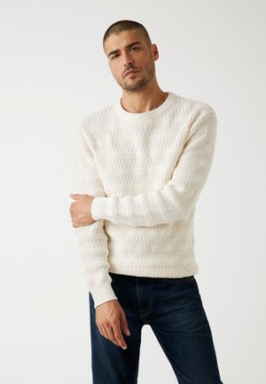 Maglione bianco lavorato a maglia con un motivo testurizzato, scollatura rotonda e polsini a costine, abbinato a jeans scuri. Il tessuto appare caldo e traspirante.