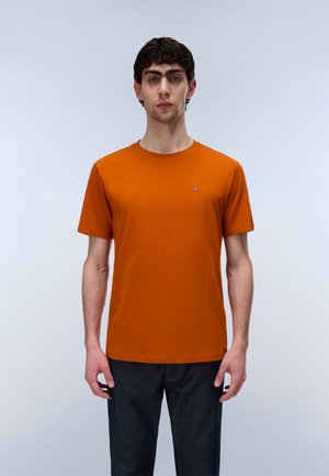 Homme aux cheveux bouclés foncés portant une chemise orange à manches courtes et un pantalon foncé, debout devant un fond clair uni.