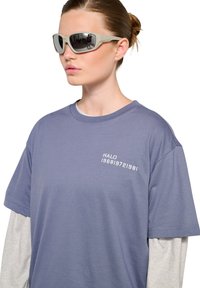 Szare okulary przeciwsłoneczne o smukłym designie i lustrzanych soczewkach. Model nosi niebieską koszulkę z białym nadrukiem oraz warstwową, szaro-długą koszulę pod spodem.