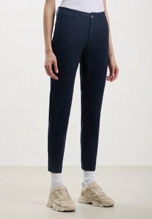 Frau trägt marineblaue Slim-Fit-Hose, weiße Socken, beige Sneakers und ein weißes ärmelloses Oberteil vor schlichtem Hintergrund.