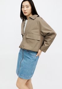 Jeune femme asiatique portant une veste beige zippée avec de grandes poches et une jupe en denim bleue longueur genoux, debout avec la main dans la poche.