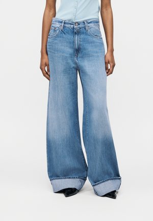 Személy világoskék, széles szárú, felhajtott szárú farmert, világoskék ujjatlan inget és fekete hegyes orrú cipőt visel, egyszínű háttér előtt áll.