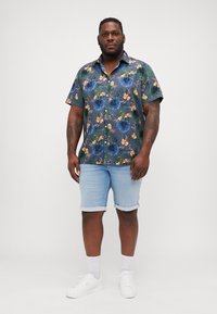 Camisa de manga corta, abotonada, con un patrón floral azul, combinada con pantalones cortos de mezclilla claros y zapatillas blancas. El fondo es de un gris liso.