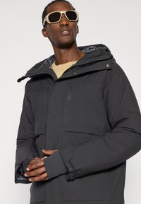 Czarna izolowana kurtka z kapturem, z dwoma przednimi kieszeniami, elastycznymi mankietami i gładką podszewką. Model w beżowej koszuli i okularach przeciwsłonecznych.