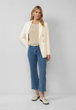 Blazer crème, pull en tricot beige, jean bleu cropped, et chaussures slip-on beiges. Boutons et textures remarquables du blazer croisé.